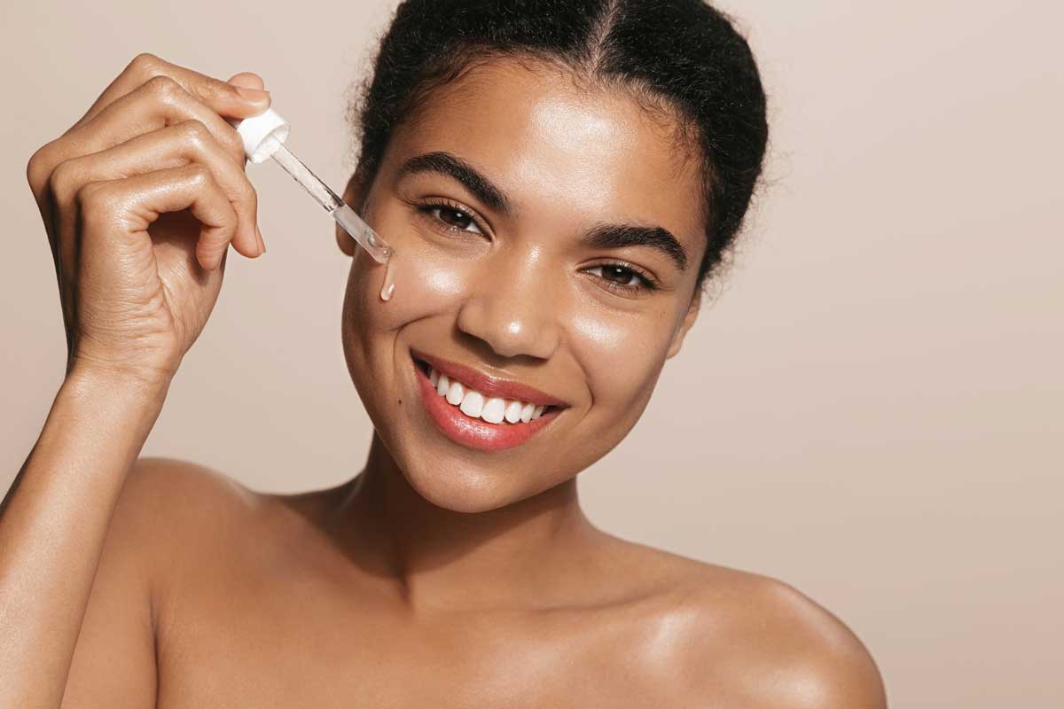 Smiling woman with perfect skin, applies tea trea, collagen face lifting serum, holds dropper, stands over brown background